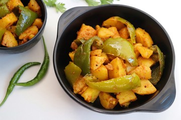 Dry capsicum and potatoes curry. also known as Aloo shimla mirchi Masala. served in black bowl with chilies and coriander over white background with copy space.