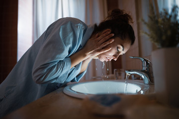 Young woman washing her face in bathroom sink.