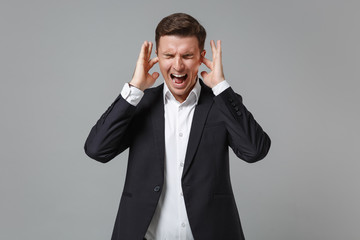 Crazy young business man in classic suit shirt isolated on grey background. Achievement career wealth business concept. Mock up copy space. Covering ears with fingers, screaming, keeping eyes closed.