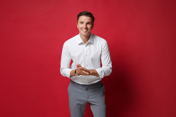 Cheerful young business man in white shirt, gray pants posing isolated on bright red wall background studio portrait. Achievement career wealth business concept. Mock up copy space. Rubbing hands.