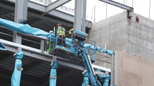 Close up of construction worker wearing protective clothing and safety harness, standing in the platform of an elevated boom lift, wrapping steel beam in a blue protective membrane