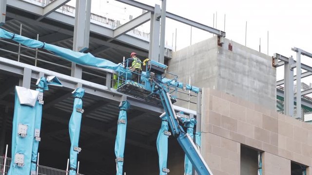 Construction worker standing on the platform of an elevated boom lift, wrapping steel beam in a blue protective membrane, on a construction site