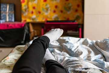 Thin female legs and feet in black pants and gray socks lie on the bed among colored fabric textiles and pillows in a well equipped room of the apartment