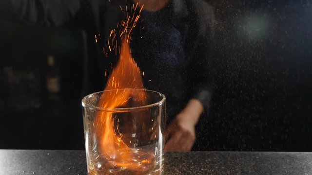 Bartender sets fire to cocktail, burning cinnamon in alcohol drink on black background. Flames in cocktail glass in slow motion, burning cinnamon in alcohol drink, barman makes drink. Full hd footage