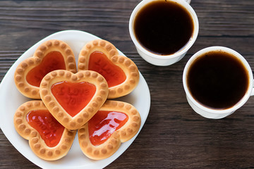 Heart-shaped cookies with strawberry jam on a white plate and two white cups of coffee on a brown wooden background. Love concept. Top view.