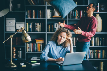 Woman working hard whit laptop while man having fun at modern home office. Man distracting the girl...