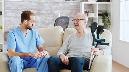 In retirement home male nurse is talking with an old disabled man with crutches next to him