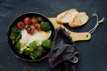 Healthy breakfast: broccoli, cherry tomatoes and fried eggs in a pan, which stands on a wooden stand on a dark background. 