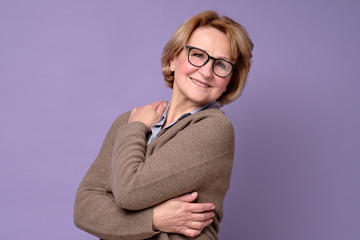Positive senior caucasian woman in glasses smiling, keeping arms around herself.