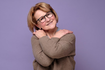 Positive senior caucasian woman in glasses smiling, keeping arms around herself.