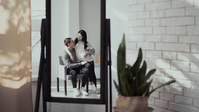 beautiful long haired pregnant wife and handsome man hug sitting together on brown chair and reflecting in large mirror at home with brick wall