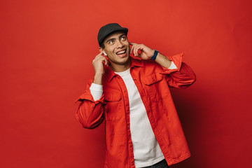 Happy positive young egyptian guy look up and smile. Listen to music through white wireless earphones. Almost dancing and having fun alone. Isolated over red background.