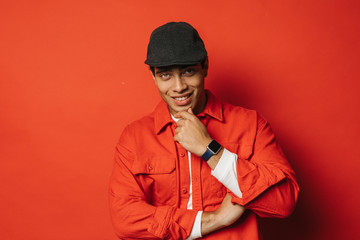 Charming, mysterious and attractive arabian or egyptian guy on picture. Touch chin with fingers and smile. Guy wear black cap and stylish clothes. Isolated over red background.