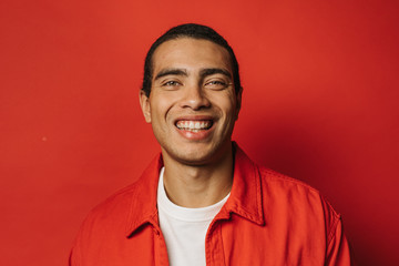 Cheerful attractive and positive young man posing on camera and smile. Nice portrait of hispanic guy. Wear colorful clothes. Cheerful smile. Isolated over red background.