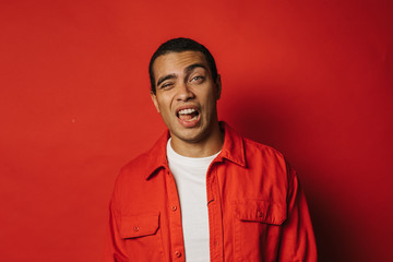 Playful young man having fun and posing on camera alone. Weird facial expressions. Emotional problems. Wear colorful clothes. Isolated over red background.