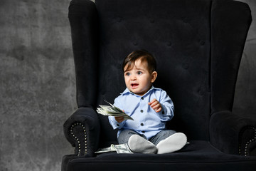 Little child in blue shirt, gray pants, white booties. He holding some money, sitting in black armchair on gray background.