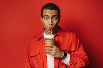 Busy young arabian man drink tea or coffee through straw. Look straight on camera and pose. Egyptian man hold paper cup in left hand. Isolated over red background.