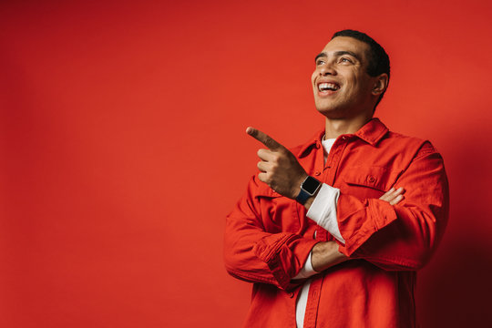 Positive happy young arabian man talking and smiling. Pointing forward. One hand is crossed. Wear colorful clothes. Posing on camera. Isolated over red background. Side view.