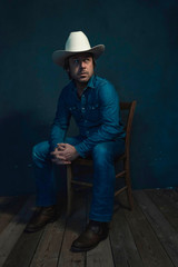 Man in jeans wearing white cowboy hat sits on a chair.