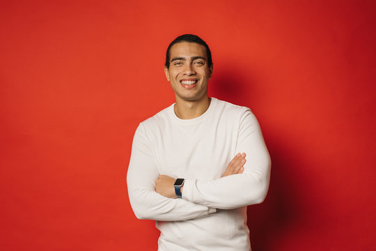 Attractive confident young arabian man posing on camera and smile. Stand with hands crossed. Wear white sweater. Egyptian or hispanic guy isolated over red background.