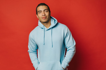 Handsome attractive young hispanic or arabian man look straight and pose on camera. Hold hands on hoodie pockets. Wear blue sweater. Stand alone isolated over red background.