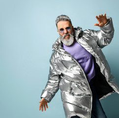 Aged male in purple sunglasses, oversized silver colored down puffy jacket, jeans. He posing on blue background. Close up