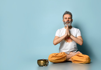 Bearded aged man in dhoti clothes. Praying, sitting on floor in lotus pose on blue background....