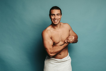 Hot sporty young arabian man show his muscles on forearms and pose on camera. Look straight and smile. White towel cover hips. Strong well-built man semi naked. Isolated over background.