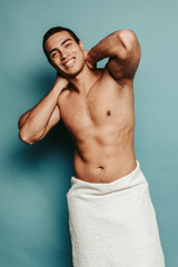 Vertical picture of young attractive man posint on camera topless. Hot sexy powerful egyptian guy hold hands behind head on neck. White towel around hips. Isolated on blue green background.