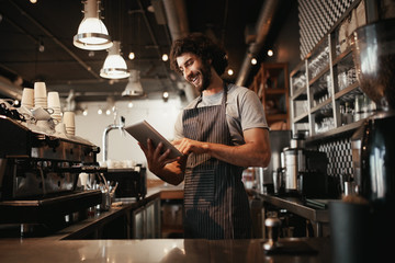Cheerful young caucasian cafe owner wearing black striped apron using digital tablet