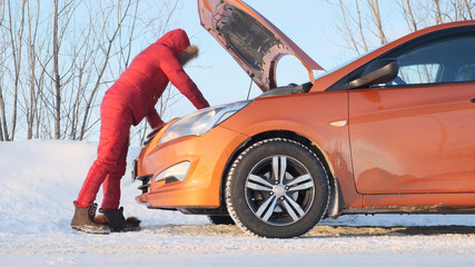 A girl in a red winter red suit stands next to a broken car.
