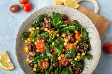 Salad with fresh crispy vegetables, chickpeas, mushrooms, spinach, cherry tomatoes, in a grey ceramic bowl, on a wooden board, blue background.
