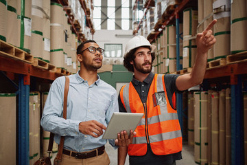 Warehouse manager wearing helmet pointing towards shelf in warehouse