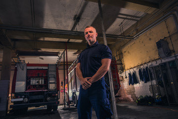 Fototapeta premium Portret przystojny strażak stojący przed ciężarówkami w remizie