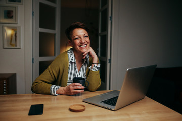 mid aged woman sitting desk at home drinking coffee and using laptop