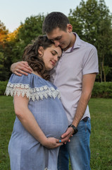 Young couple in love on a walk in the park, husband hugs pregnant wife by the belly, happy family