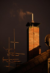 A bricked chimney with smoke together with some antennas on the roof of a house in the last rays of...