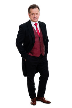 Full lenght Portrait of handsome confident caucasian mature businessman 55-60 years old in black tuxedo and red vest standing with hands in pockets smiling in front on isolated white background.