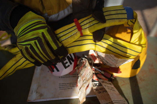Top view of rigger inspector high risk worker hand wearing heavy duty glove holding inspecting a red safety tag on yellow three lifting sling defocused personal safety lock pre inspecting book helmet 