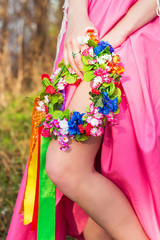 Colorful wreath with ribbons in woman's hand and pretty woman's leg seen from bright pink dress. The celebration of the pagan Slavic holiday of Ivan Kupala Day or Midsummer. Slavic national traditions