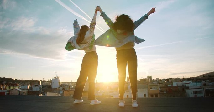 Shot from back of two female silhouettes happy raising hands up outdoor on rooftop sunset, joyful young girl friends enjoying Barcelona city view of old town and sun on background dressed in jackets