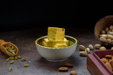Soan papdi dessert cubes in bowl, cardamom grains in wooden scoop, pistachios and almond on concrete kitchen surface with black copy space.