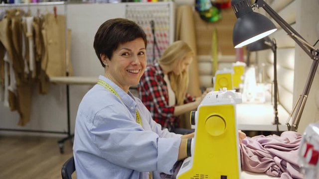 Tilt down medium shot of smiling middle aged seamstress with short hair looking at camera while using sewing machine sitting at table in studio, her young colleague working in background