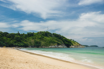 Fototapeta na wymiar Phuket, Thailand - April 19, 2017. Sea beach with people and umbrellas, a large green hill and a wind generator.