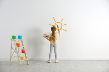 Little child painting sun on white wall indoors