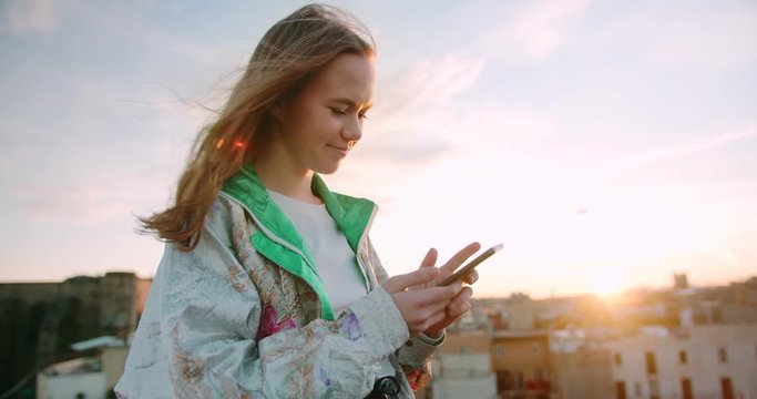 Portrait of happy cute teen Caucasian girl using mobile device to be online in app outdoor, young nice ginger hipster female standing at rooftop enjoying sunny urban moment sharing in social media