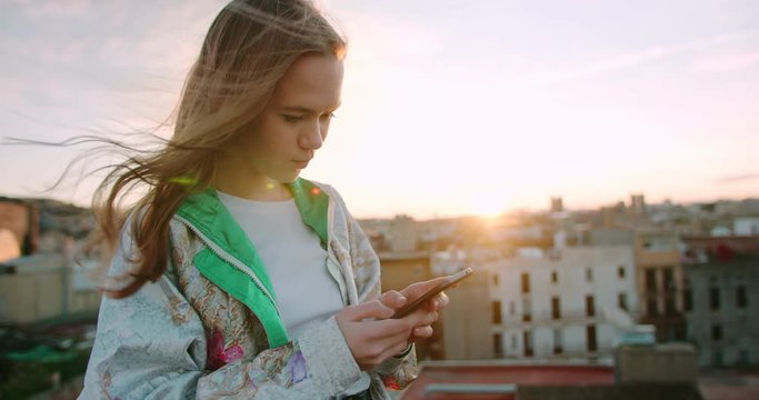 Cheerful Caucasian young female chatting in new messenger application using smartphone, attractive ginger teen girl using smartphone device outside at rooftop with old city view and sunset bokeh