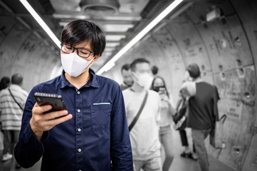 Asian man wearing surgical face mask using smartphone in subway tunnel with crowded people walking. Wuhan coronavirus (COVID-19) outbreak prevention in public area. Health care and medical concept