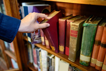 Male university student hand choosing and picking vintage book from old wooden bookshelf in college...