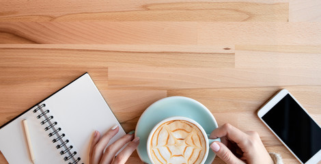 Aroma coffee in woman hands with mobile phone and  notebook with pencil on a wooden background with copy space. Morning cappuccino in cafe and relax. Top view and banner.